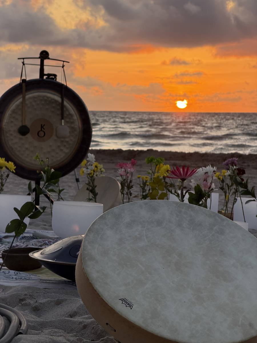 A gong silhouetted against the setting sun, with flowers and a frame drum on the sand